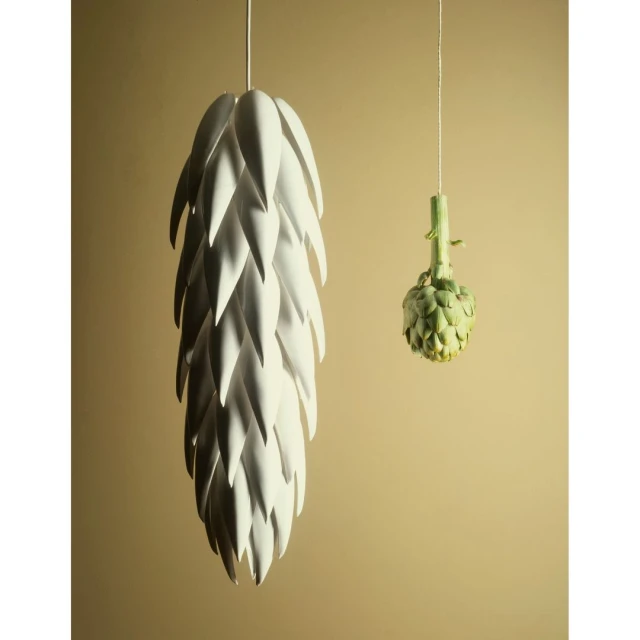 Artichoke-shaped pendant lamp alongside an actual artichoke hanging against a tan background in a studio setting. Interior design.