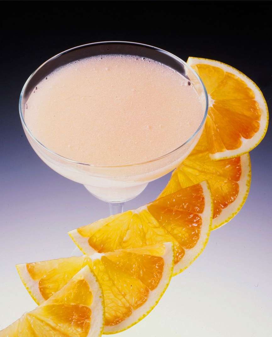A cocktail glass filled with a creamy orange drink next to a curved line of sliced oranges against a light gradient background.