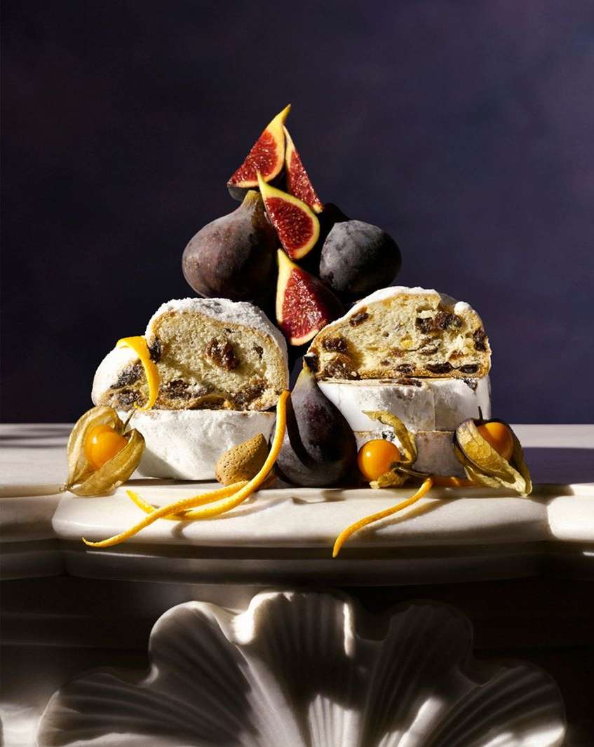 Overhead food photo of a stollen cake sliced open with figs, almonds, and kumquats on a marble surface against a dark background.