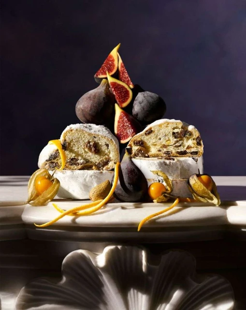 Overhead food photo of a stollen cake sliced open with figs, almonds, and kumquats on a marble surface against a dark background.