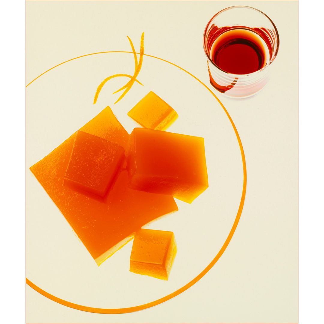 Orange gelatin dessert cubes on a clear plate next to a clear glass with a red liquid, minimalist food still life photography.
