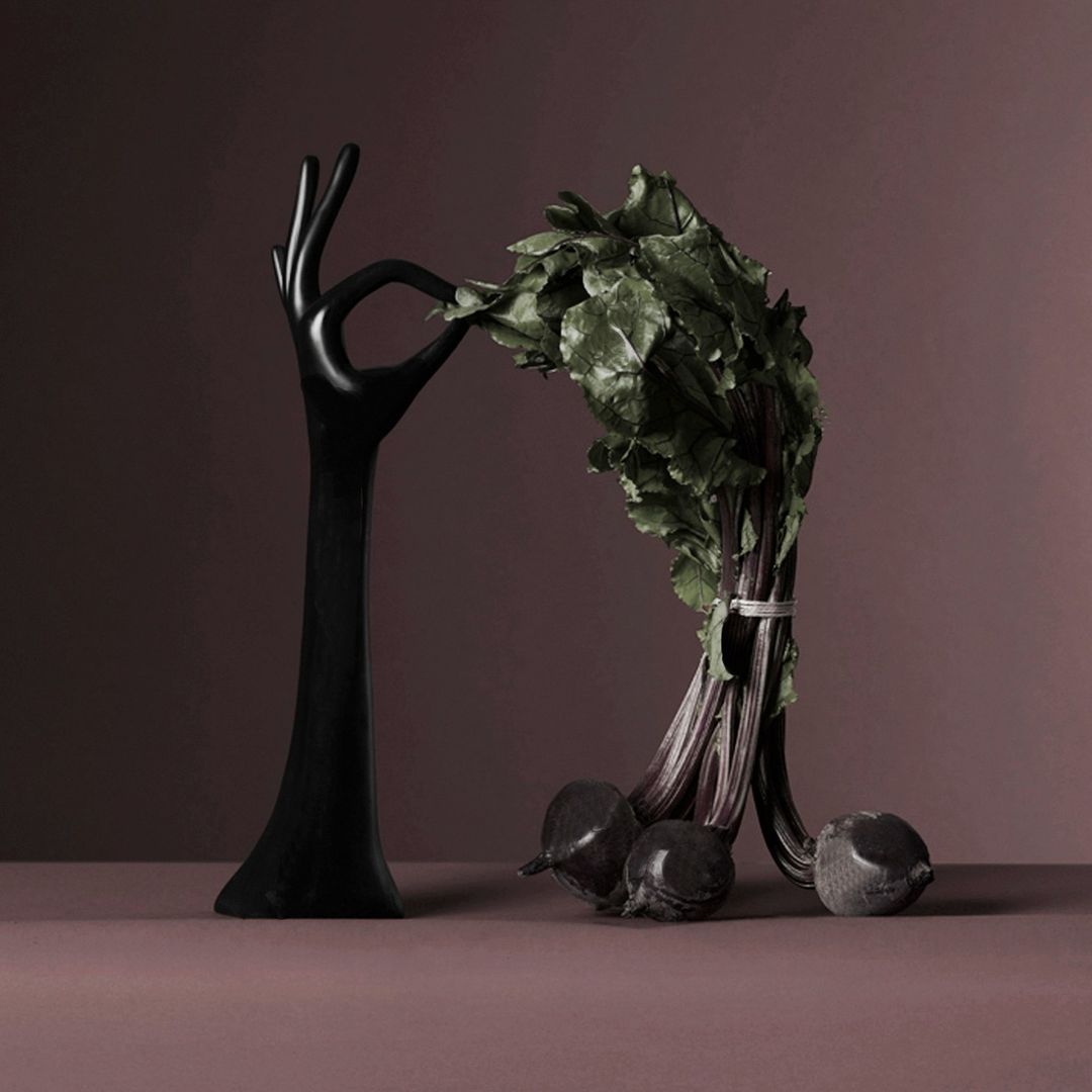 Still life of a black, hand-shaped sculpture holding a bunch of beets with green leaves against a dark purple background.