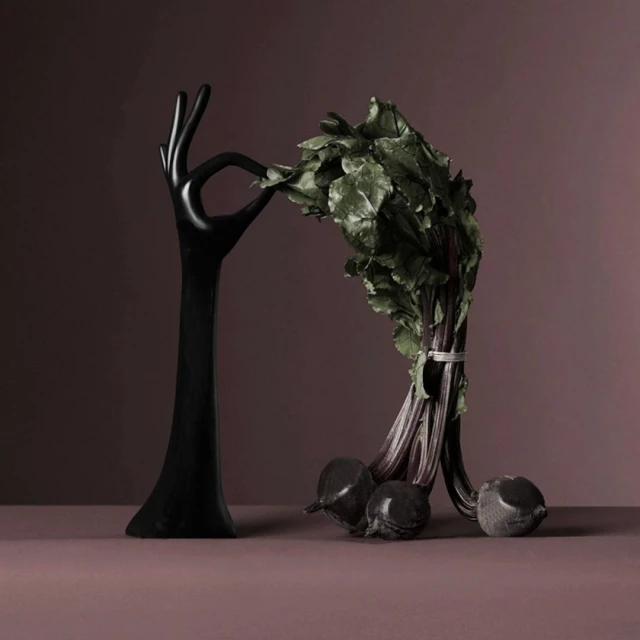 Still life of a black, hand-shaped sculpture holding a bunch of beets with green leaves against a dark purple background.