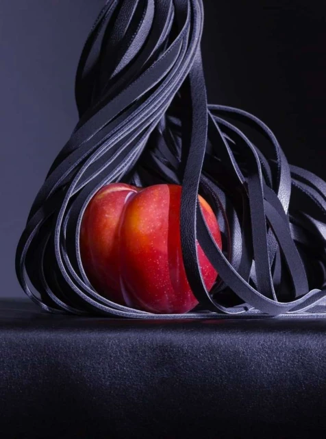 Dramatic studio shot featuring a bright red plum nestled among looping black leather straps on a dark surface with an almost black backdrop.