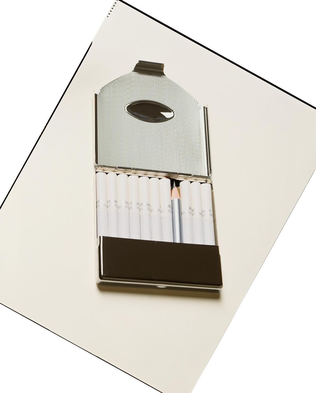 Open silver cigarette case with white cigarettes and a single white pencil on a white surface, angled shot, indoor studio.