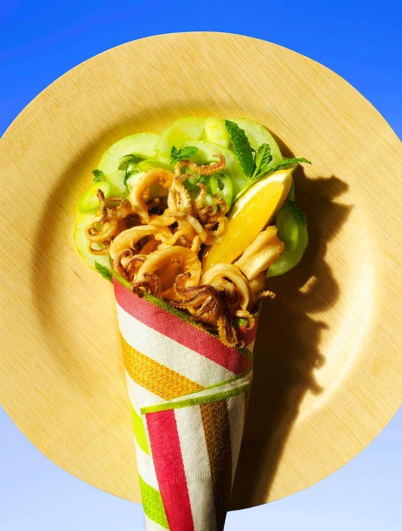 Overhead shot of fried calamari, cucumber slices, and lemon on a wooden plate, served in a colorful striped paper cone against a blue background.