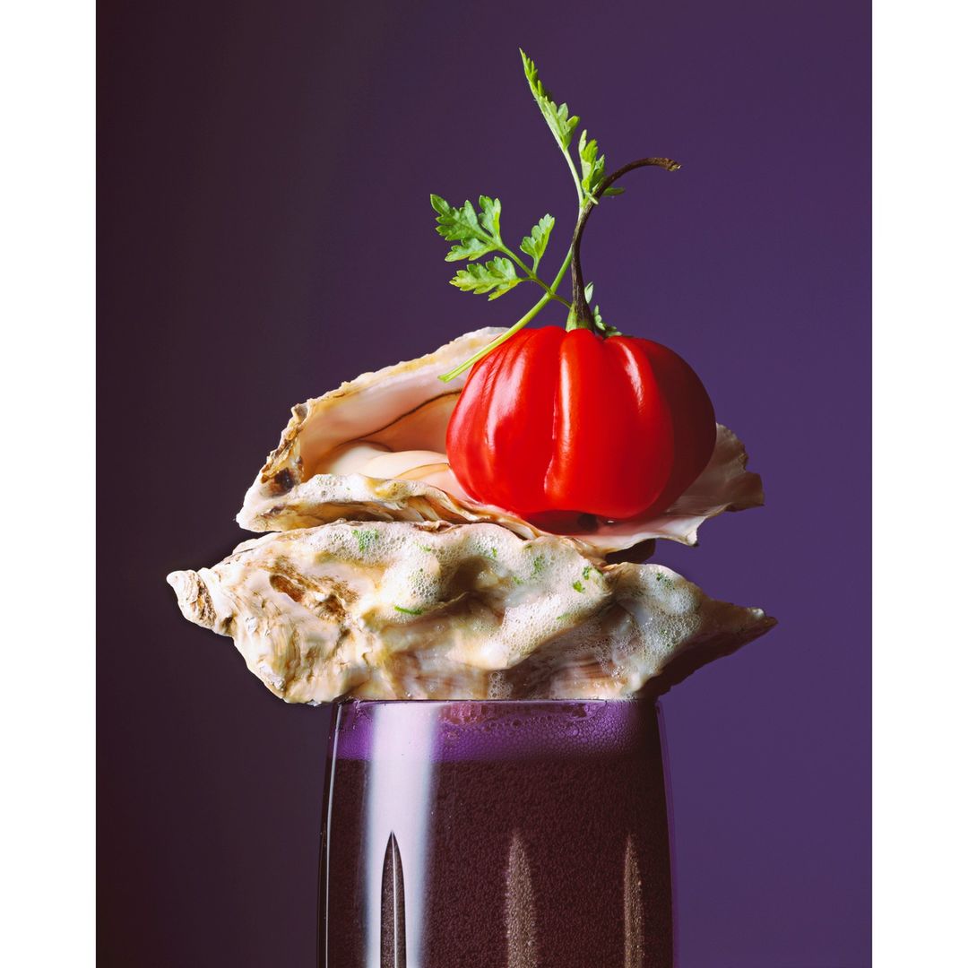 Food photography of a red tomato and parsley sprig sitting on top of oyster shells and a dark purple drink against a dark purple background.