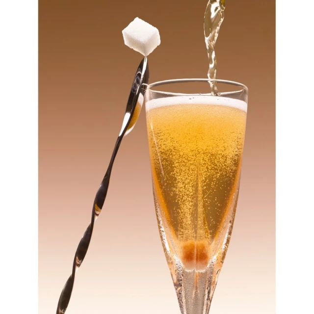 Champagne flute filled with bubbling champagne with a sugar cube on a twisted silver spoon. Light amber background.