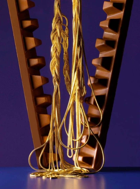 Golden chain necklaces draped on a dark brown, geometric stand against a blue backdrop, showcasing texture, reflection, and modern jewellery.