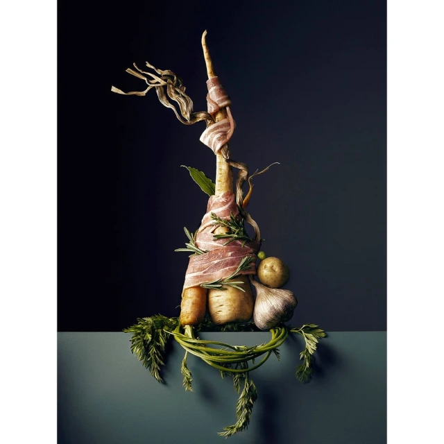 Still life food photo with parsnip wrapped in bacon, garlic, potatoes and herbs against a dark gray background on a gray surface.