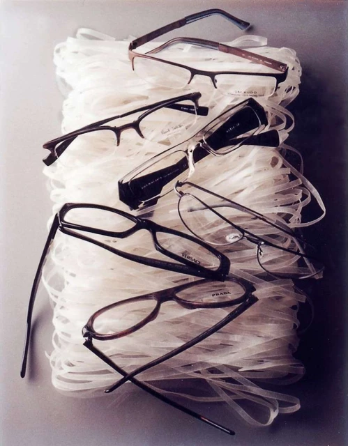 A pile of designer eyeglasses including Versace and Prada brands are stacked on top of a base of shredded, translucent white material.