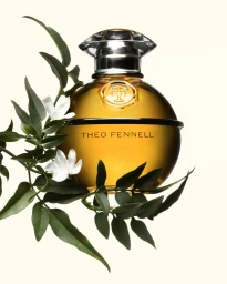 A glass perfume bottle with amber liquid and white flowers and green leaves framing the base against a light background.