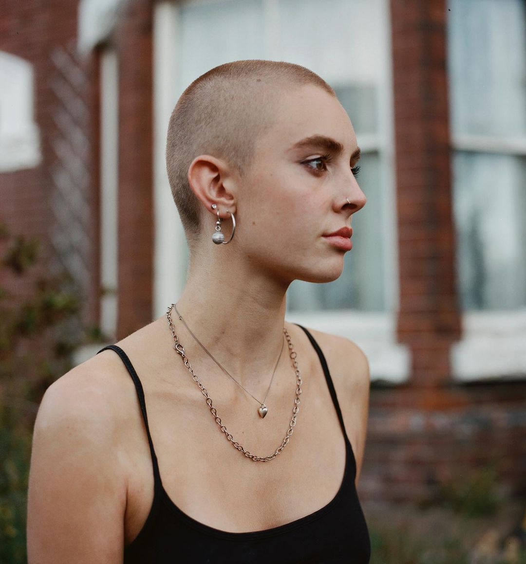 Portrait of a young adult female with a shaved head, wearing a black tank top, a silver hoop earring, and necklaces, posing outdoors.