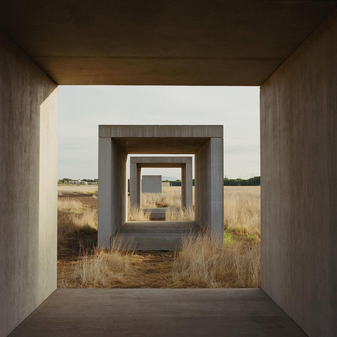 Concrete cube structures create a tunnel effect through a field of dry grass toward the horizon under a light cloudscape.
