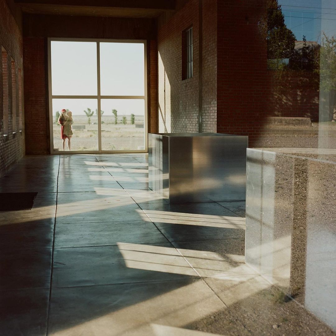 Woman and baby standing by large window in industrial space; shadows and reflections on concrete floor, brick walls, desert landscape.