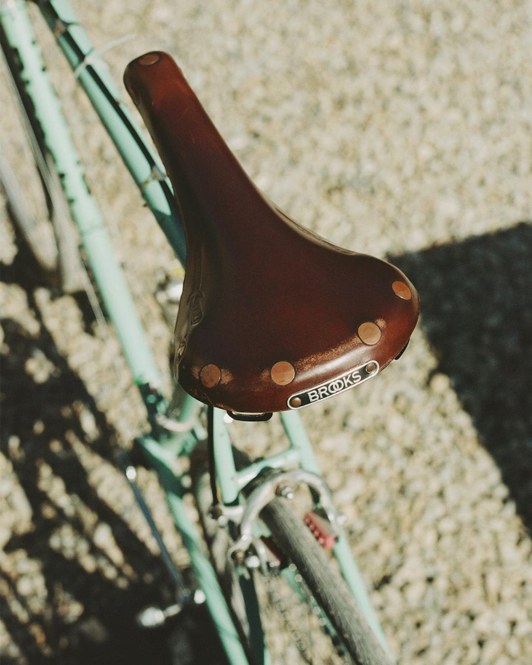 Close-up on a light blue bicycle featuring a brown leather Brooks saddle with copper rivets, set against a neutral gravel background.