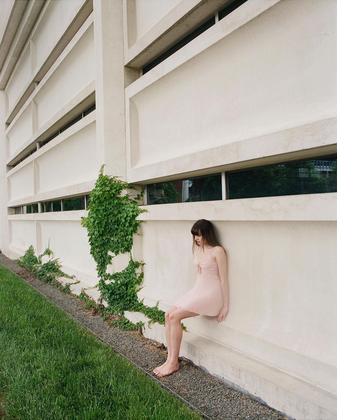 Young woman in a pink dress sits barefoot on a ledge of a modern building, lush green ivy climbs the white facade, natural light.