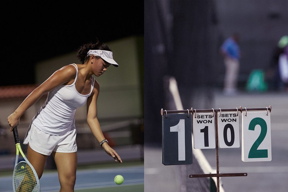Young woman in white tennis outfit serves with racquet on blue court; scoreboard with '1' and '2' shows match in progress, nighttime.