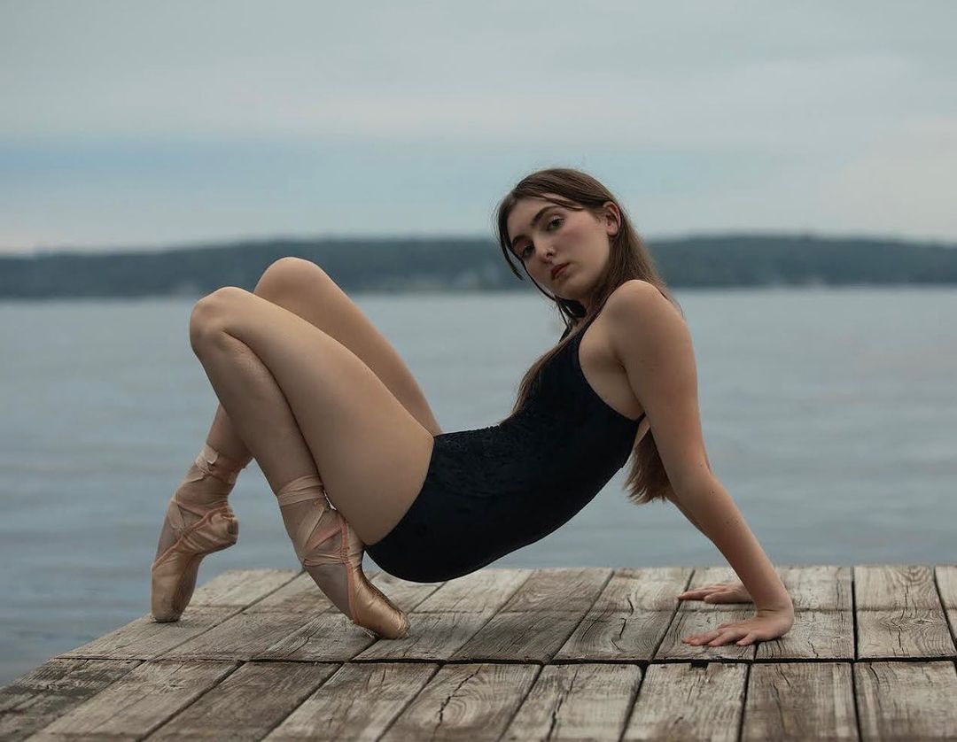 Female ballet dancer in black leotard and pointe shoes poses on a wooden dock near the water, looking at the camera with a neutral expression.