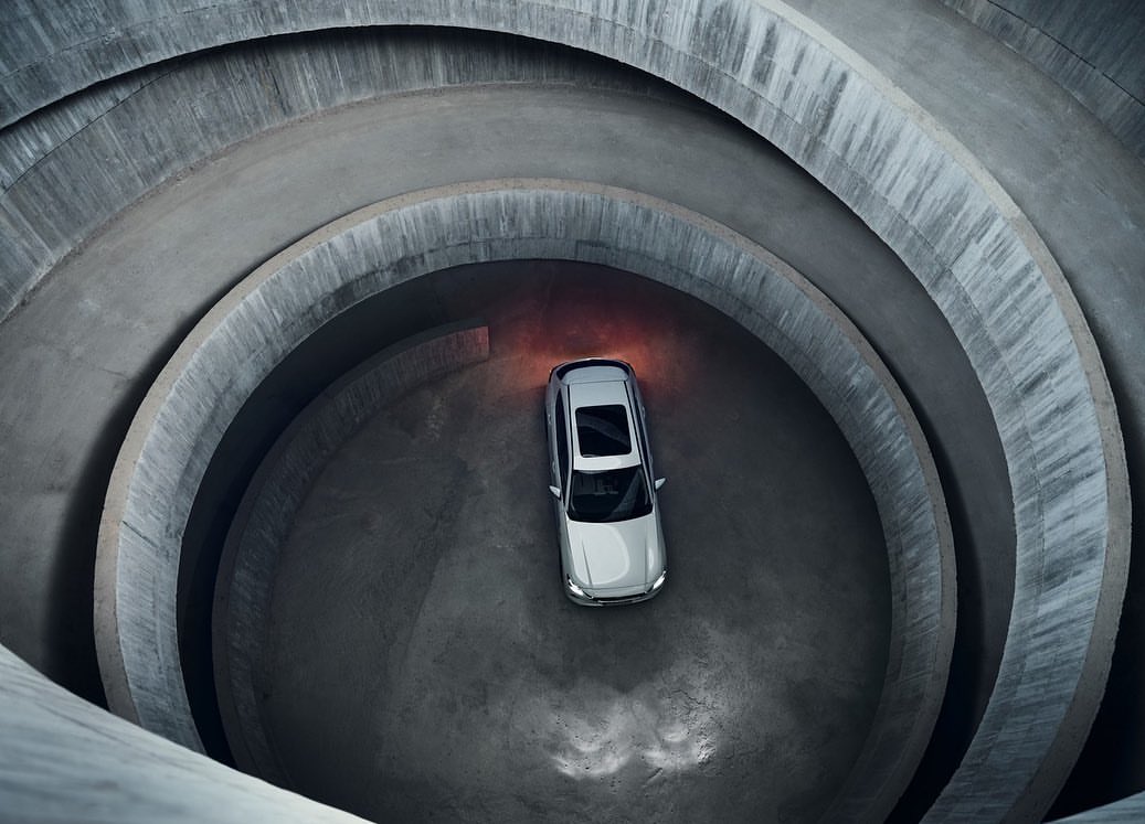 Aerial view of a modern silver car driving through a concrete parking garage ramp with sunlight entering the structure.