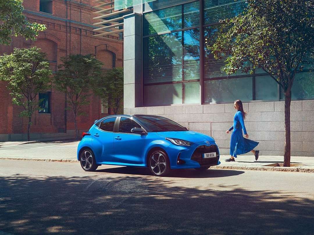 Blue Toyota Yaris parked on a city street with a woman walking in the background, blending modern architecture and nature.