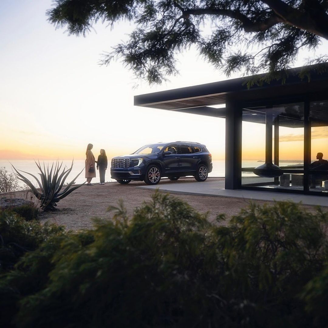 A dark blue SUV sits parked near a modern house while an adult female and a child look out over the ocean at sunset.