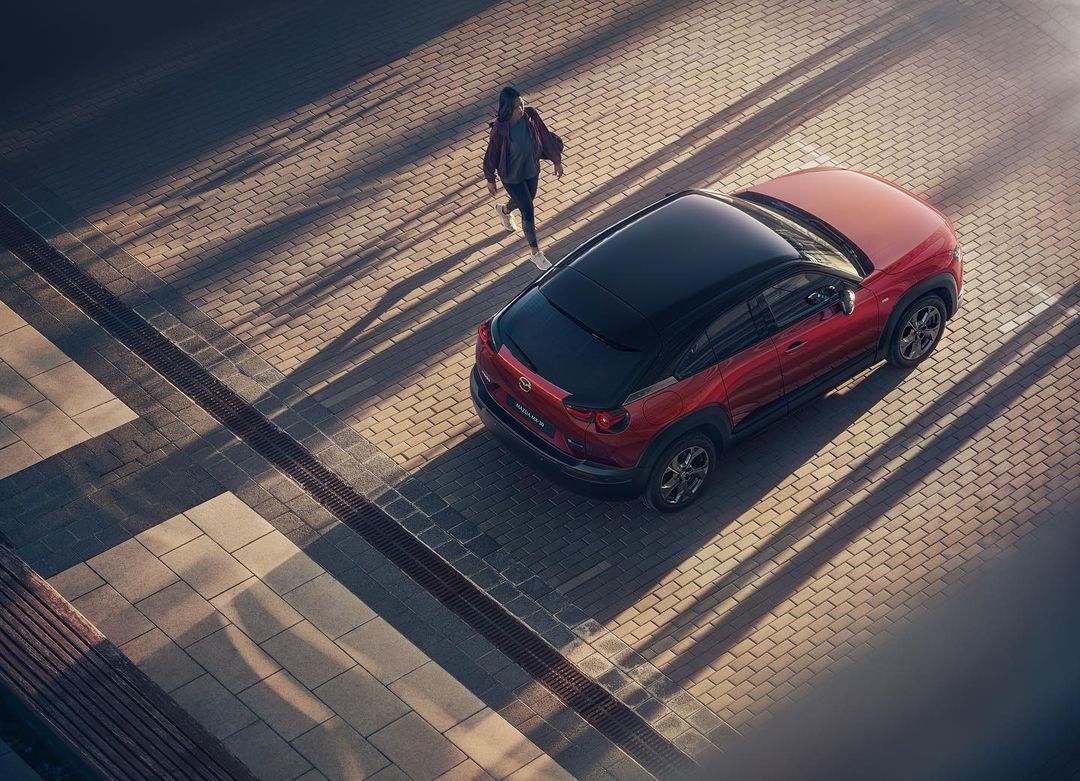 Overhead shot of red Mazda MX-30 parked on brick street with person walking nearby, illuminated by sunlight creating long shadows.