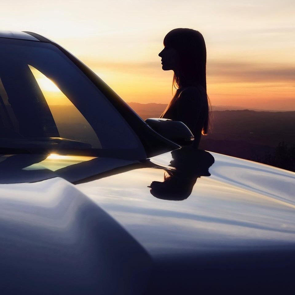 Silhouette of a woman standing near a car at sunset with soft, warm lighting, and a distant mountain range.