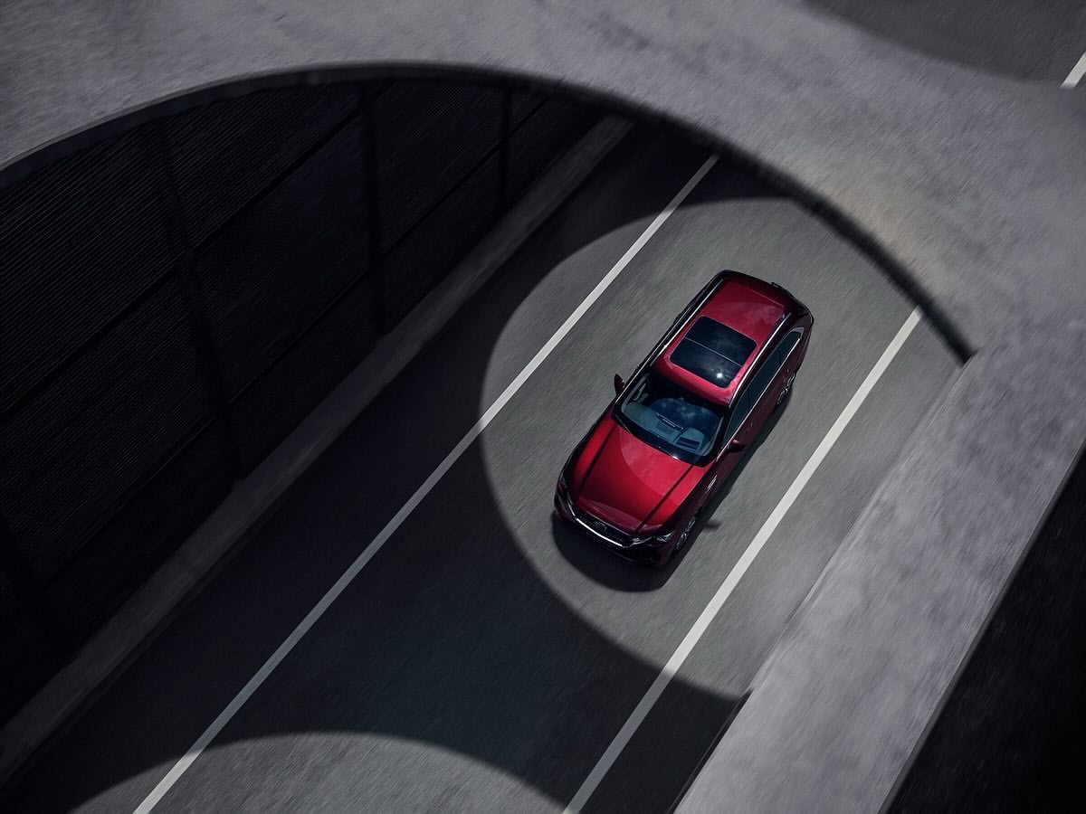 Aerial shot of a red sedan driving on an asphalt road, framed by overhead concrete structures creating a tunnel-like effect.