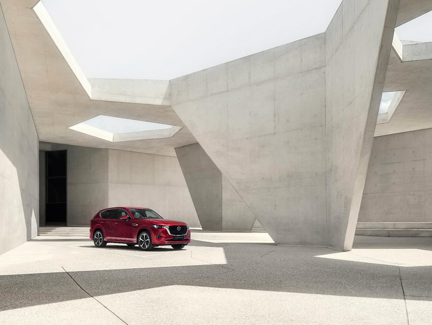 Red Mazda CX-50 SUV parked in front of a modern, light gray concrete building featuring geometric shapes and a bright, open space.