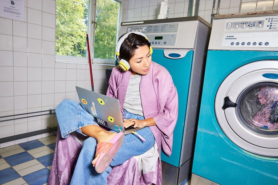 Woman sitting on a stool in a laundromat with a laptop and headphones next to washing machines, illuminated by natural window light.
