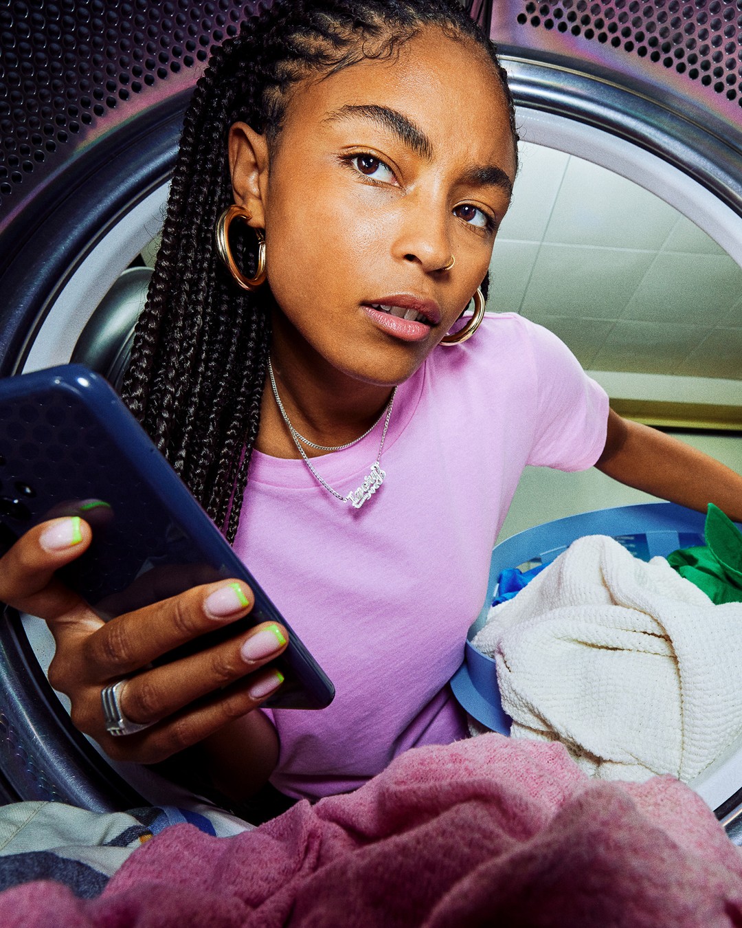 A woman with braided hair holds a cell phone while standing next to a washing machine filled with clothes, illuminated by soft light.