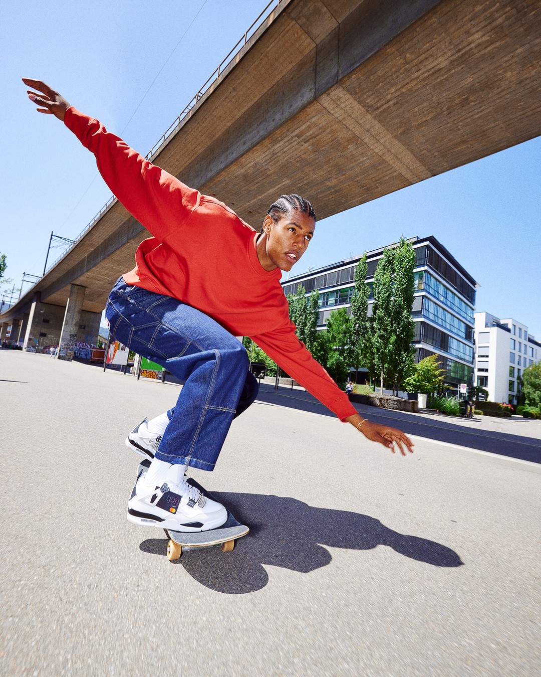 A young man in a red shirt and blue jeans is skateboarding on an asphalt surface on a sunny day with a building and bridge in the background.