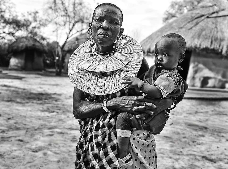 Black and white portrait of a Maasai woman in traditional attire holding a young child, captured outdoors with simple dwellings in the backdrop.