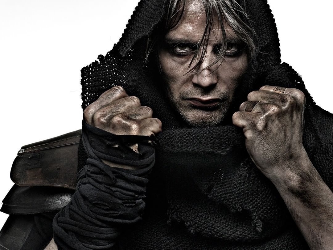 Close-up of a gritty adult male with hooded headwear and clenched fists, set against a bright white background in a photo studio.