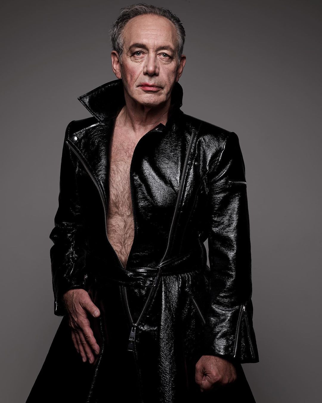 Studio portrait of a mature man in a black patent leather coat with an open front, posing against a neutral gray background.