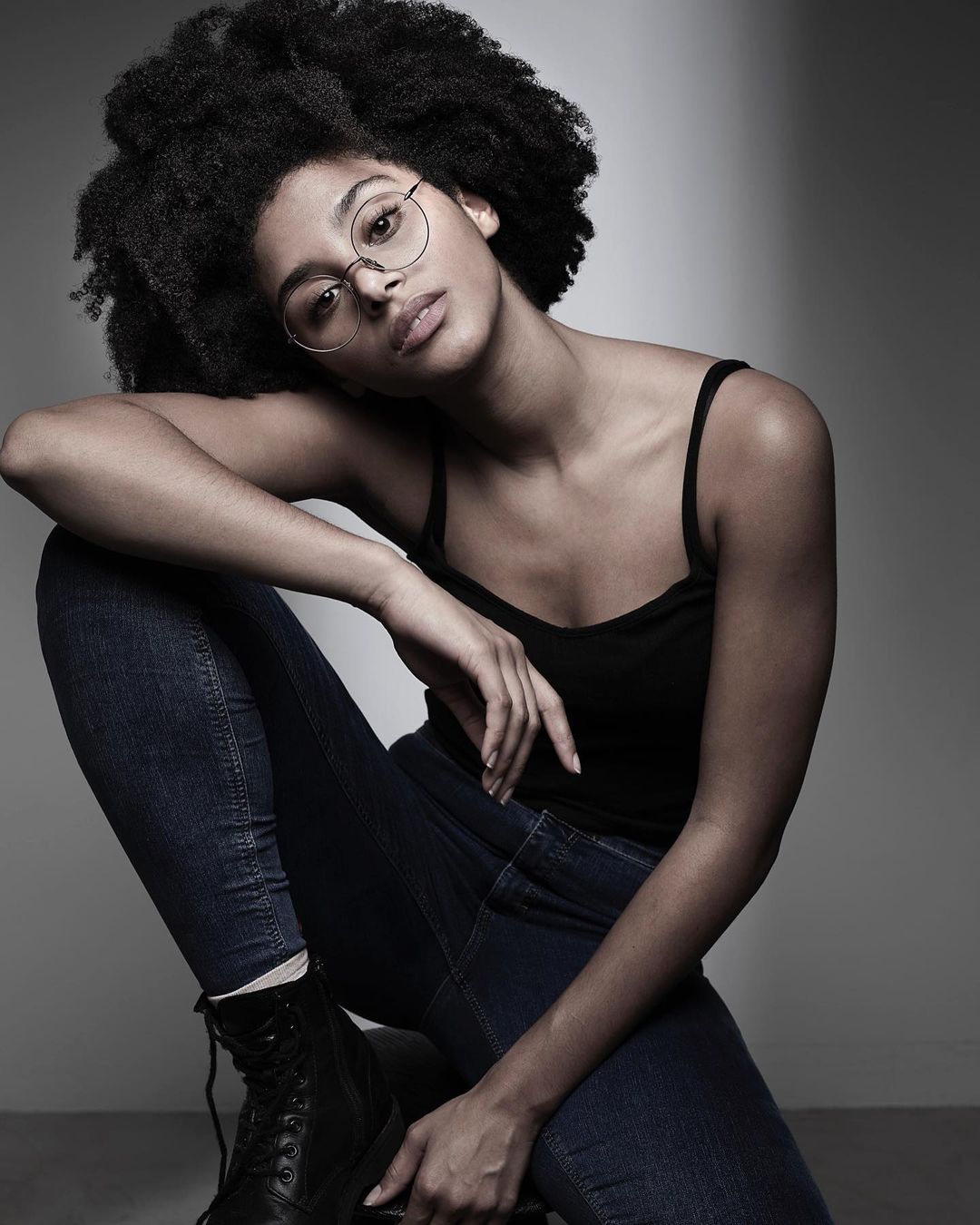 Portrait of a young woman with dark curly hair, wearing a black tank top, blue jeans, round eyeglasses and boots, posing in a photo studio.