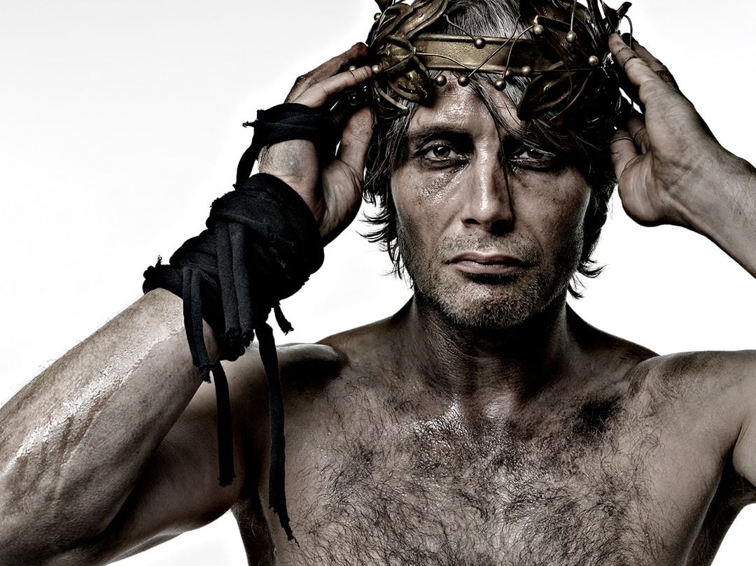 Portrait of a muscular adult man with a laurel crown, smudged makeup, and black armbands in a studio setting on a white background.