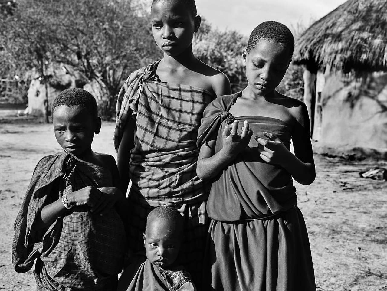 Black and white shot of four young children in traditional clothing, posing outdoors with a thatched-roof hut in the background on a bright day.
