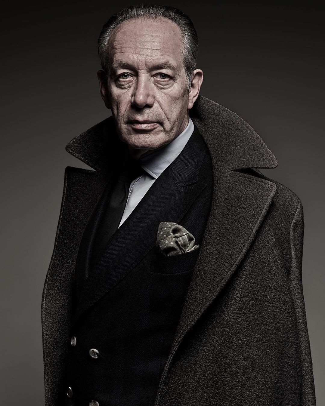 Portrait of an older man in an overcoat and suit, looking directly at the camera with a serious expression in a photo studio.