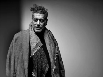 Portrait of a man with curly hair wearing a dark coat and patterned scarf against a plain studio background.