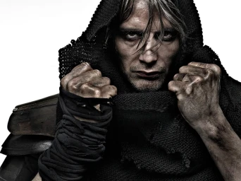 Close-up portrait of Mads Mikkelsen wearing a black hooded garment with wrapped fabric on his forearms against a white background.