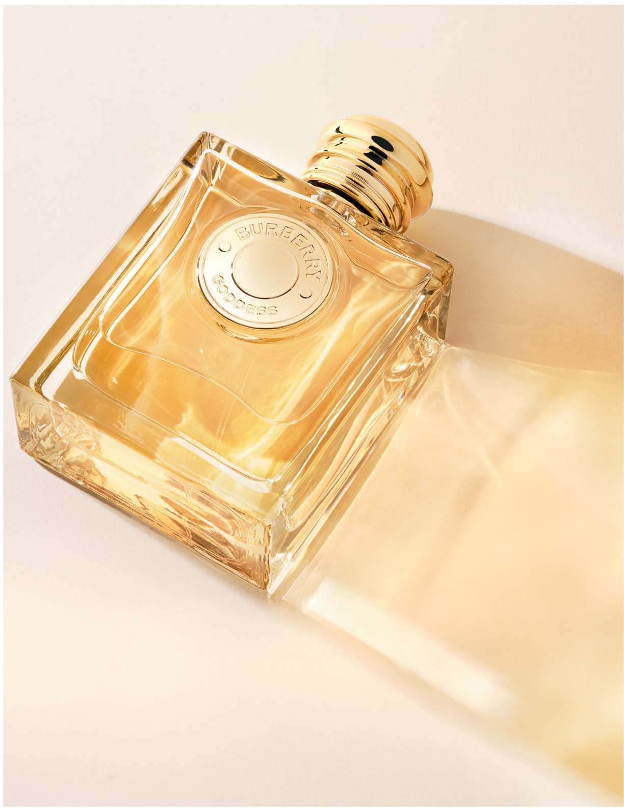 Burberry Goddess perfume bottle on its side with a transparent package, on a cream backdrop, studio lighting, and warm tones.