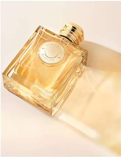 Burberry Goddess perfume bottle on its side with a transparent package, on a cream backdrop, studio lighting, and warm tones.