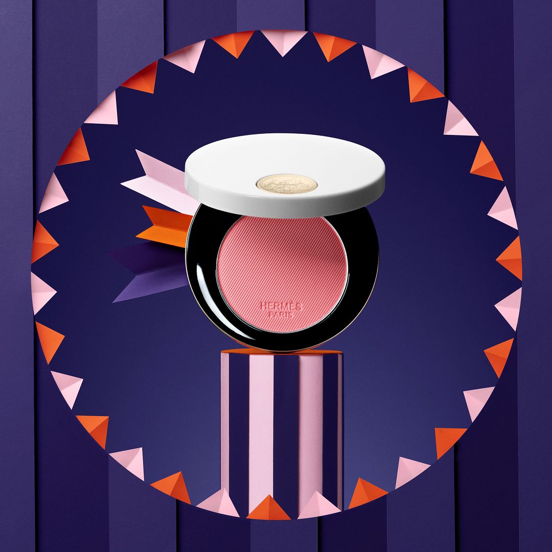 Hermès blush compact displayed on a striped stand with a colorful geometric backdrop; product shot for the beauty industry.