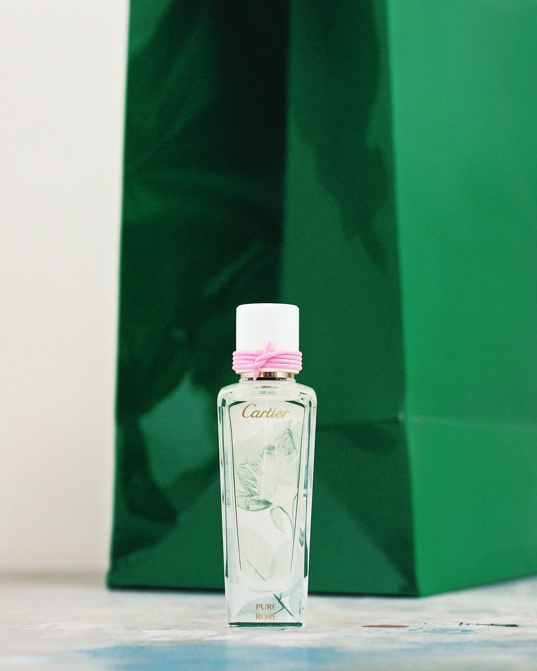 Close-up of a glass Cartier perfume bottle with a white cap and pink ribbon against a green shopping bag backdrop on a light surface.