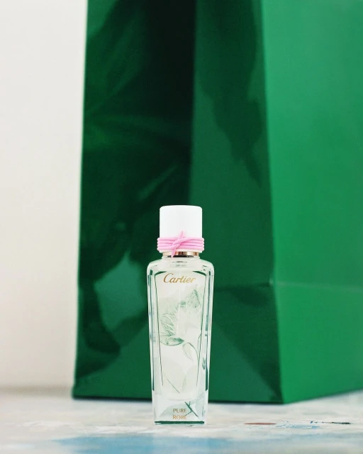 Close-up of a glass Cartier perfume bottle with a white cap and pink ribbon against a green shopping bag backdrop on a light surface.