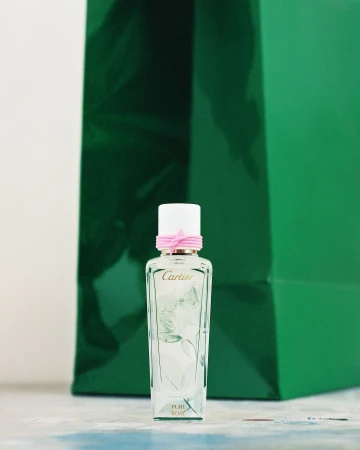 Close-up of a glass Cartier perfume bottle with a white cap and pink ribbon against a green shopping bag backdrop on a light surface.