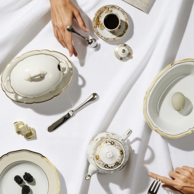 Overhead shot of elegant porcelain tableware, including teapot, plates, and serving dishes, arranged on a white tablecloth with silver cutlery.