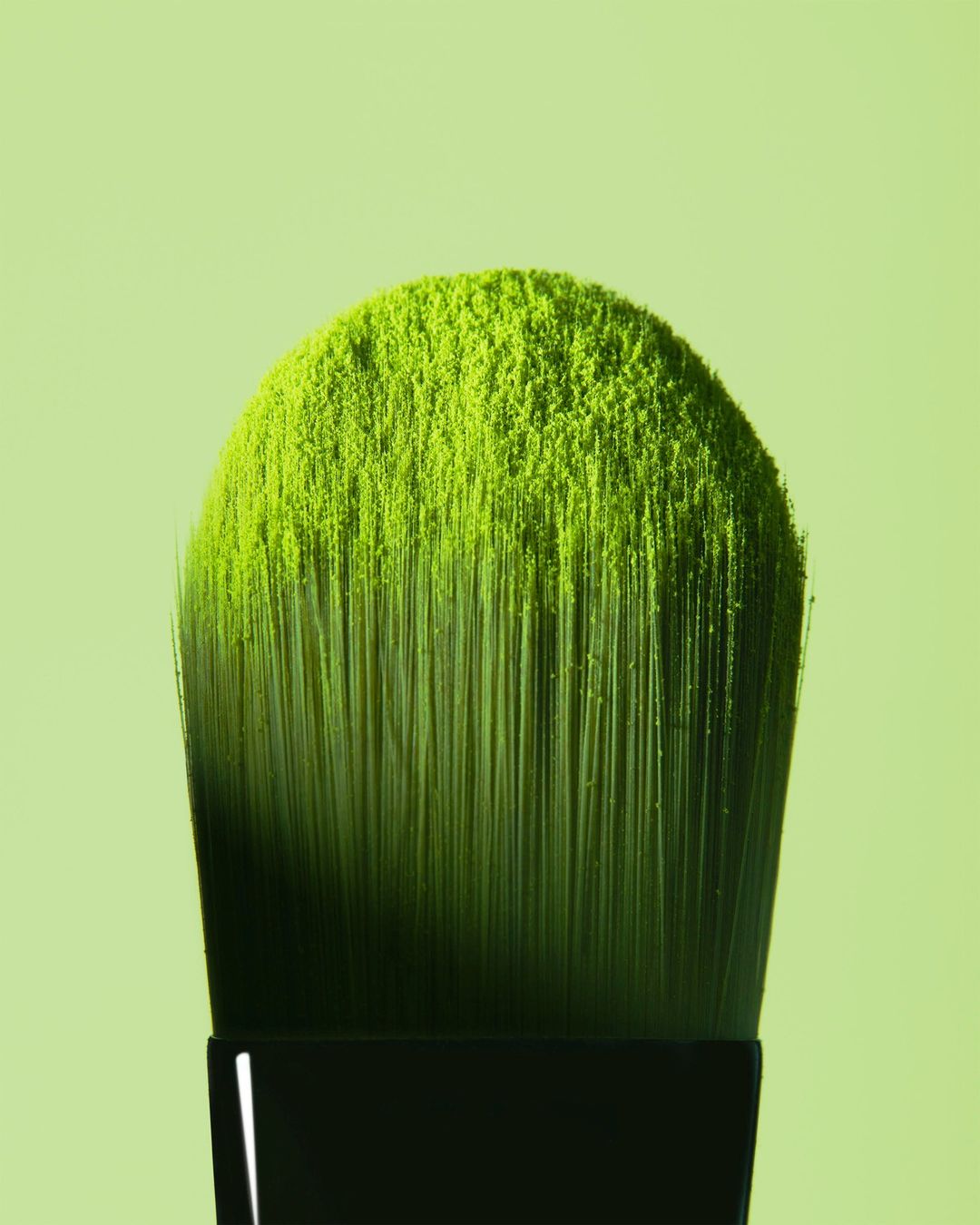 Close-up of a makeup brush densely coated with bright green powder against a soft green background, beauty concept for cosmetic product.